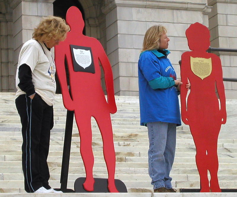 Two people standing on the Alberta Legislature for the Silent Witness project, with two red wooden figures.